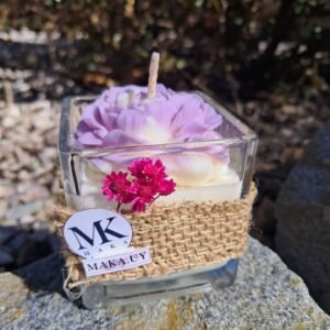Vela aromática de cera de soja en envase de vidrio cuadrado con faja de yute, flores secas fucsias y diseño de flor lila en relieve de la marca MAKA Uruguay.
