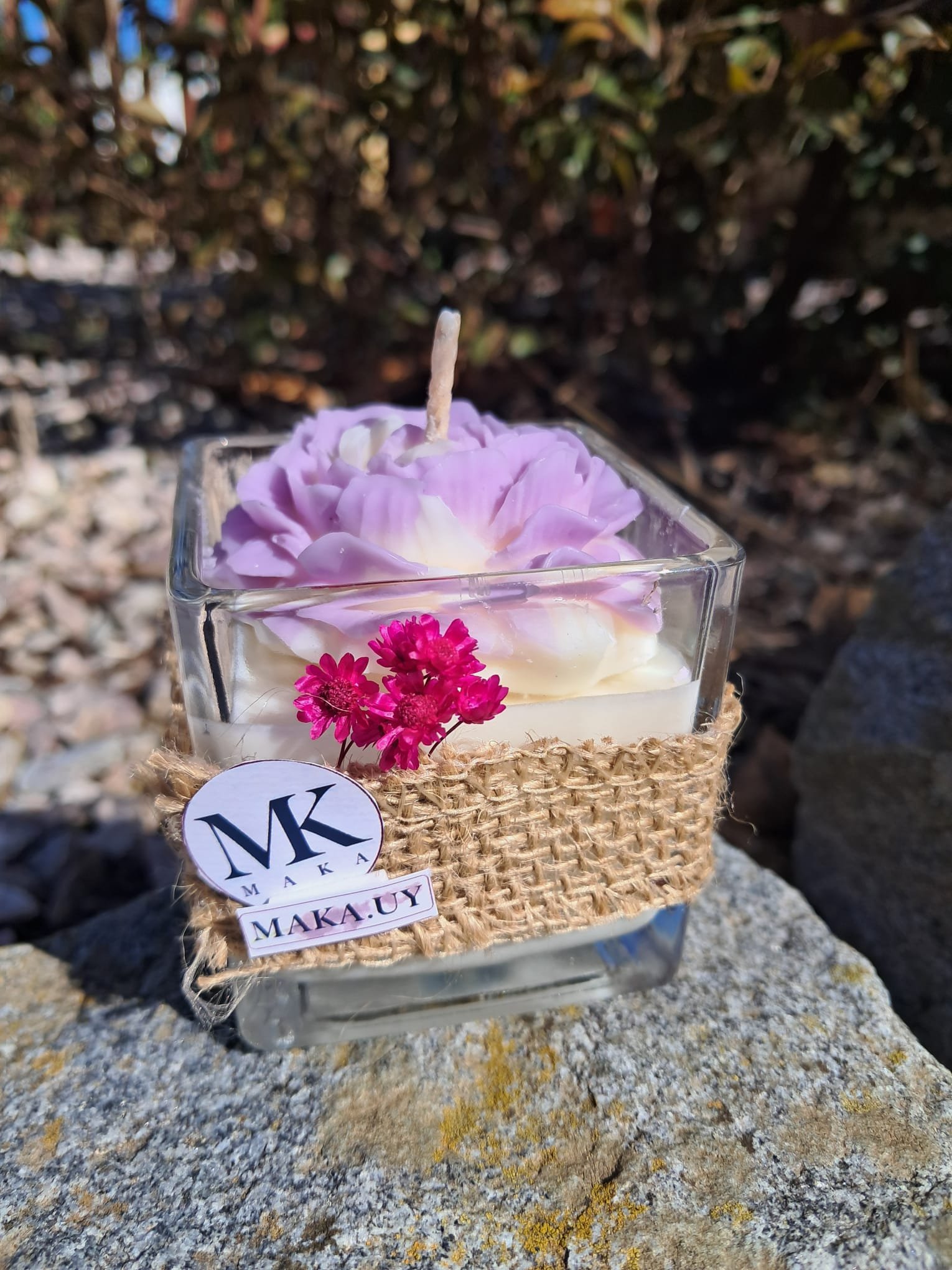 Vela aromática de cera de soja en envase de vidrio cuadrado con faja de yute, flores secas fucsias y diseño de flor lila en relieve de la marca MAKA Uruguay.
